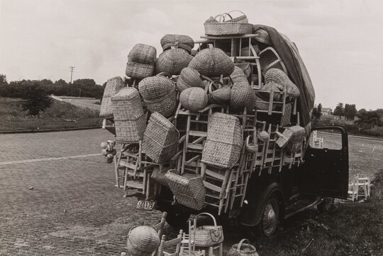 A black-and-white photograph of the back of a truck overflowing with wooden chairs and baskets.