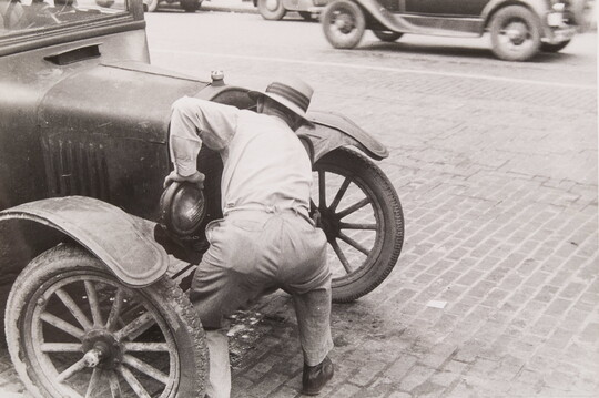 A black-and-white photograph of a man bent over the front of an early 20th-century car.