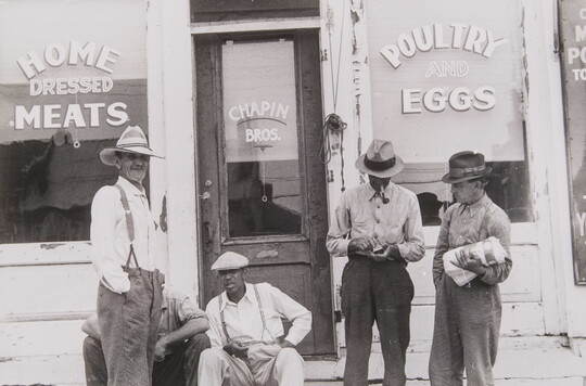 A black-and-white photograph of a group of men, all dressed in work clothes and wearing hats, sitting and standing in front of a store advertising meat, poultry, and eggs.