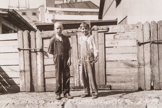 A black-and-white photograph of two White boys dressed in baggy overalls standing in front of a wood fence.