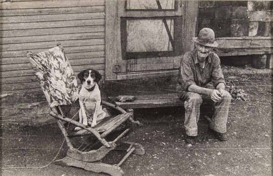 A black-and-white photograph of an elderly man in ragged clothing sitting on a wood stoop as a dog sits in a wood rocker next to him.