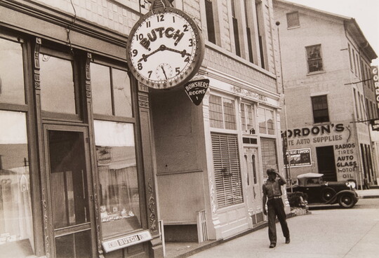 A black-and-white photograph of a Black man walking down a street lined with store fronts, one of which has a large clock face in the foreground.