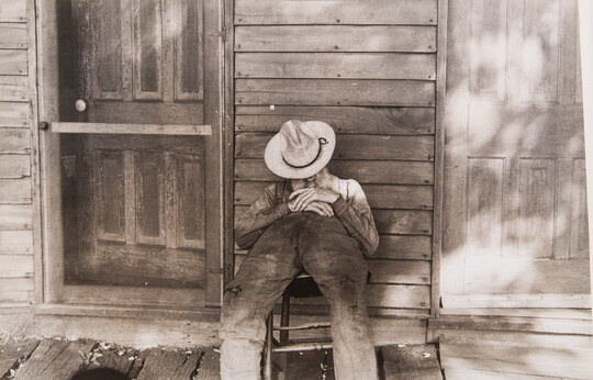 A black-and-white photograph of a man sleeping in a chair, a hat pulled over his face, on the porch of a wood-framed home.