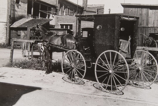 A black-and-white photograph of a horse-drawn carriage, horse tethered to a hitching post, a row of wood buildings in the background.