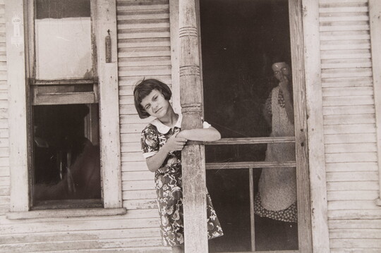 A black-and-white photograph of a young White girl leaning on a porch post as a White woman in a dress and apron stands just inside a screen door.