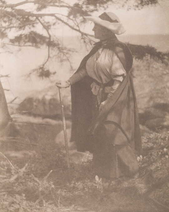 A sepia-toned photograph of a White woman walking in the woods with a walking stick, a large hat, and a cloak.