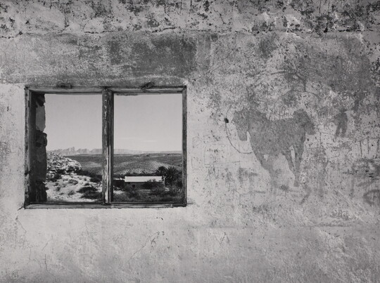 A black-and-white photograph of a concrete wall with a window that reflects an open landscape with mountains in the distance.