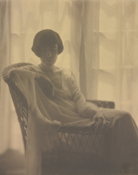 A sepia-toned portrait photograph of a White woman with a bob haircut, wearing a flowing dress and seated in a wicker chair, backlit by sun coming through gauzy curtains.