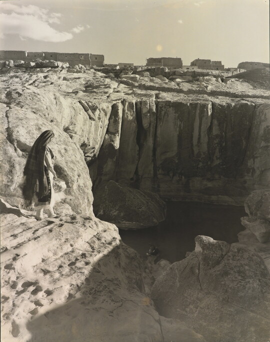 A black-and-white photograph of a woman with a long headscarf on a cliff, overlooking a water hole at the bottom.