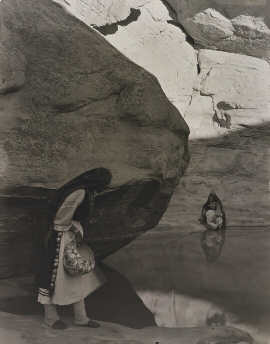 A black-and-white photograph of a woman with a long headscarf standing at the edge of a water hole surrounded by boulders and rock walls.