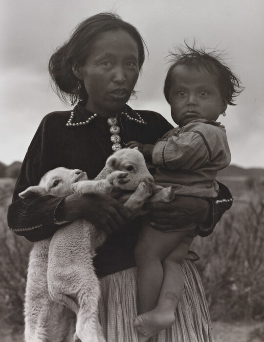 A black-and-white portrait photograph of a Native American woman holding a toddler on one hip and two lambs in her other arm.