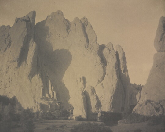 A sepia-toned photograph of tall and broad rock formations with scrubby vegetation at the base of the rocks.