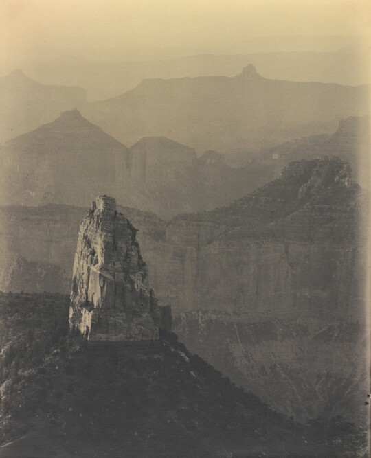 A sepia-toned photograph of rock walls and cliffs fading into the distance; a single rock tower and scrubby vegetation in the foreground.
