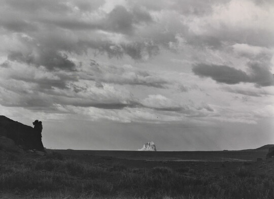 A black-and-white photograph of an open field with a large, white rock formation in the distance under a cloudy sky.