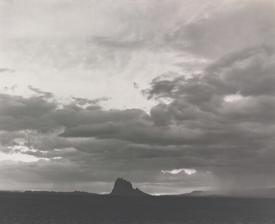 A black-and-white photograph of a dramatic light and dark sky with some striated clouds and a rock formation in the distance.