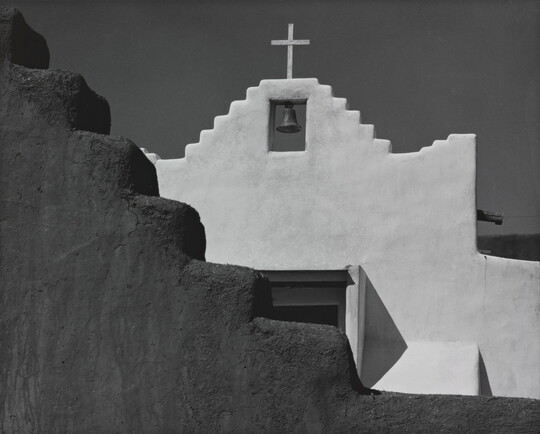 A black-and-white photograph of a terraced adobe bell tower with a cross on top, partially hidden by a terraced adobe wall.