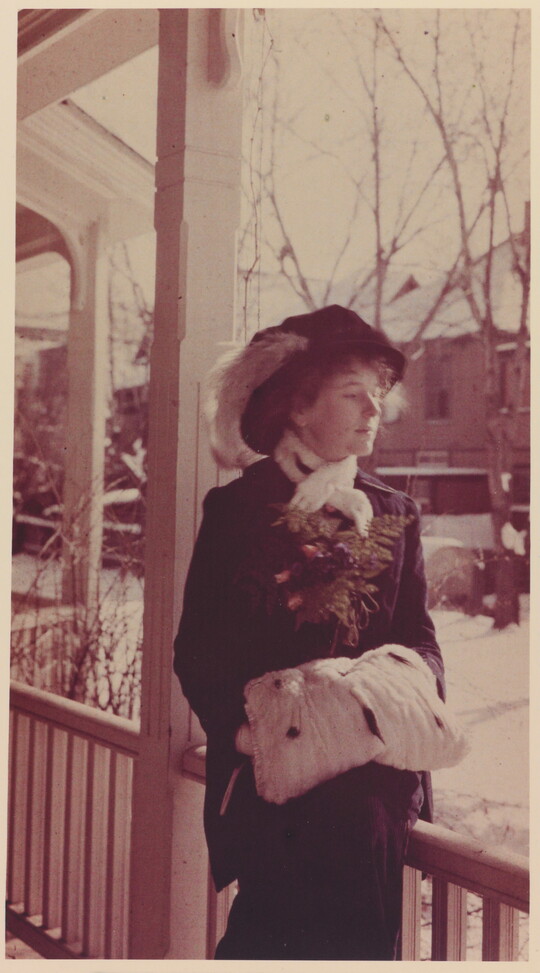 A sepia-toned portrait photograph of a White woman wearing a hat with a feather in it, a scarf, coat, hands tucked into a muff, standing on a porch.