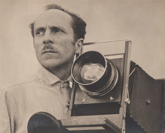 A sepia-toned portrait photograph of a balding White man with a mustache beside a large-format camera.