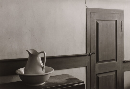 A black-and-white photograph of a ceramic pitcher and wash basin on a table next to a wooden door.