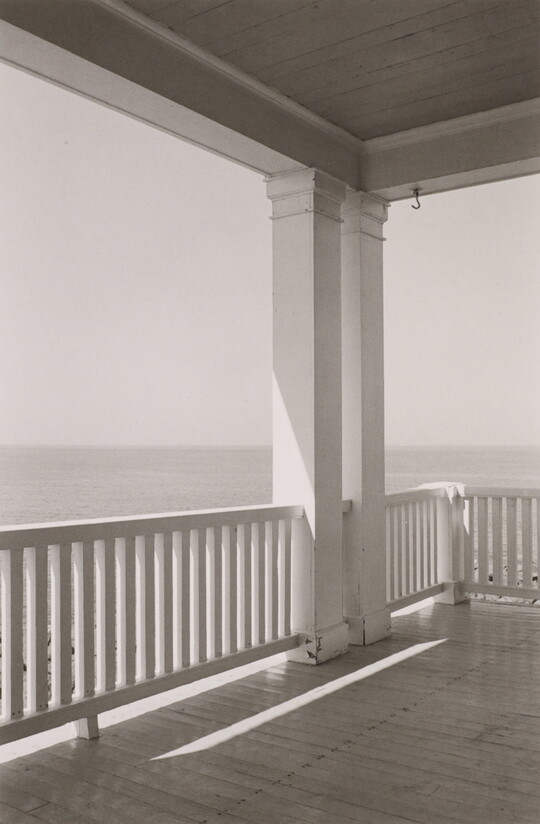 A black-and-white photograph of porch with a white-slatted railing overlooking the ocean.