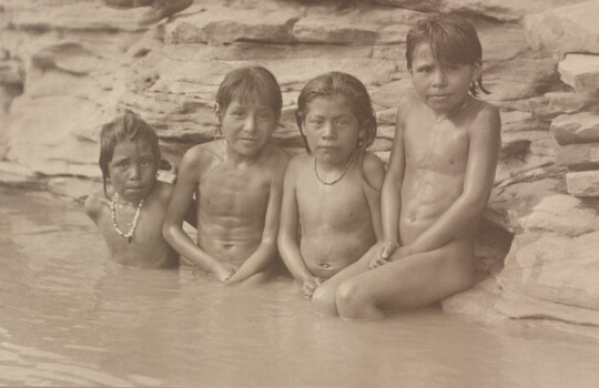 A black-and-white photograph of four Indigenous children sitting in a pond or stream leaning against its rocky edge.