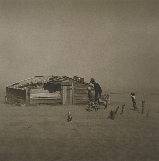 A black-and-white photograph of a man and two children walking through a dust storm by a dilapidated structure.