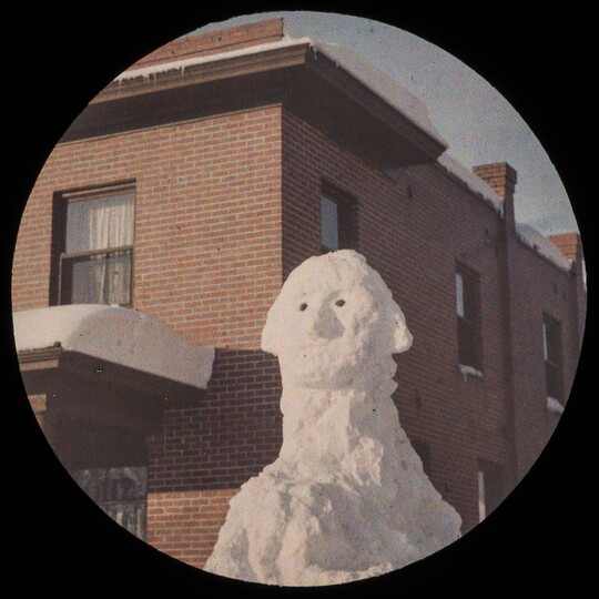 A circular color image of a cartoonish likeness of George Washington sculpted from snow in front of a red brick building.