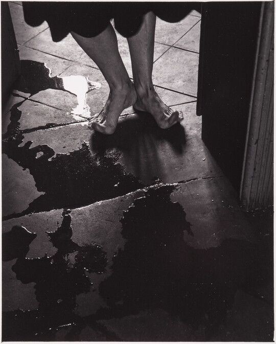 A black-and-white photograph of a person's bare feet, toes stretched up off the tile floor to avoid puddles.