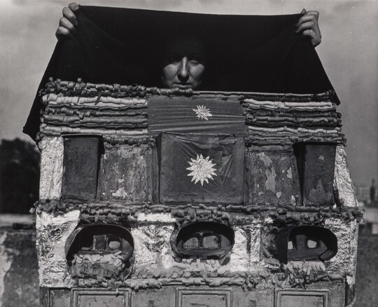 A black-and-white photograph of a person holding a black cloth above what looks like a homemade dollhouse, but instead of windows, it has stereoscopes embedded in it.