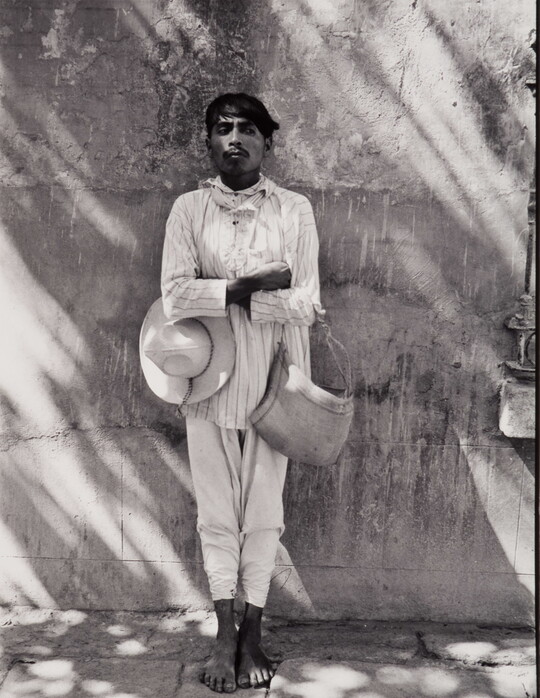 A black-and-white photograph of a dark-skinned man in light-colored shirt and pants, holding a hat under one crossed arm and a bag on the other, leaning against a wall.