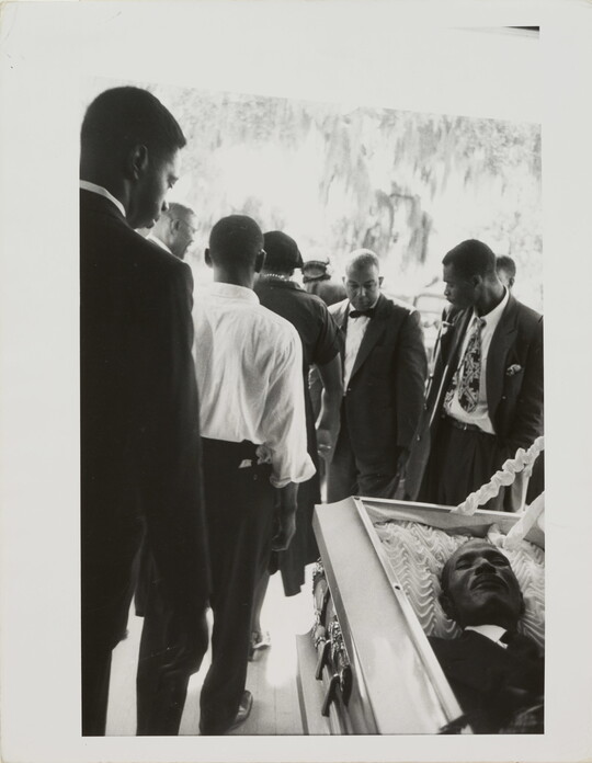 A black-and-white photograph of Black men in suits filing by an open casket.