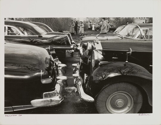 A black-and-white photograph of 1950s-style cars parked bumper to bumper; people in overcoats gather in the background.