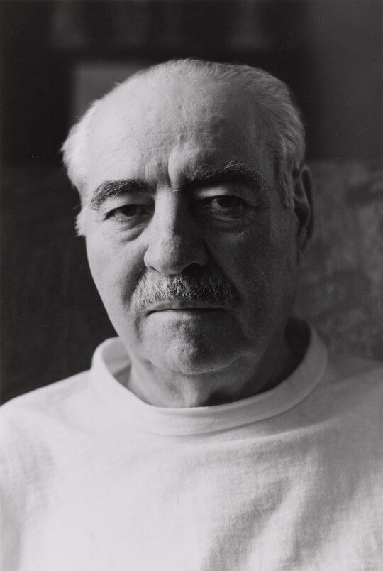 A black-and-white portrait photograph of an older, balding White man with dark eyebrows, a mustache, wearing a light shirt.