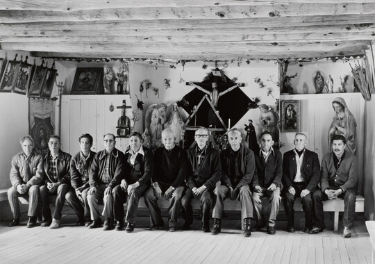 A black-and-white photograph of a group of men, most of them older, seated on a wood bench in front of many religious sculptures and paintings.
