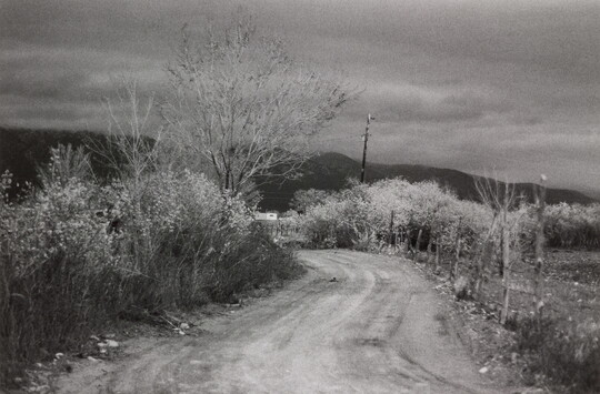 A black-and-white photograph of a curvy dirt road with scrubby vegetation on both sides, distant hills, all under a dark sky.