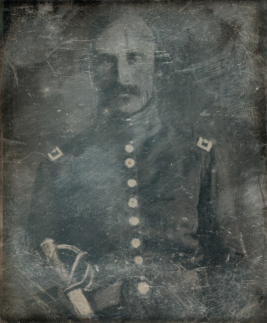 A dark, heavily scratched black-and-white photograph of a White man with a moustache, wearing military regalia and a sword.