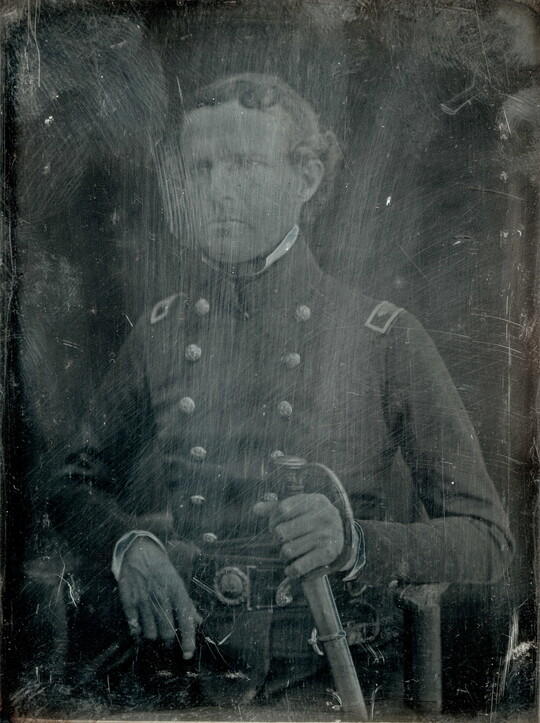 A black-and-white portrait photograph of a seated White man dressed in military uniform and holding a sword, obscured by marks and scrapes on the photograph surface.