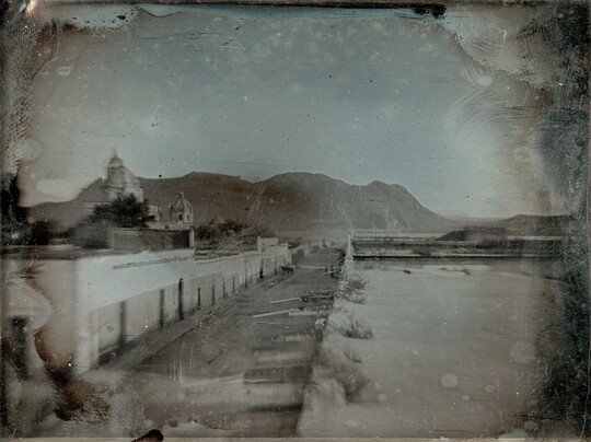 A black-and-white photograph, slightly obscured by surface marks, of a deserted street, a church, and mountain in the background.