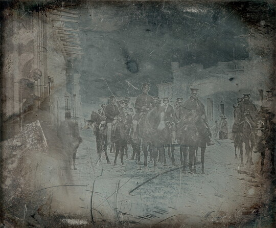 A dark, black-and-white photograph of a group of figures in military uniform on horseback on a street.