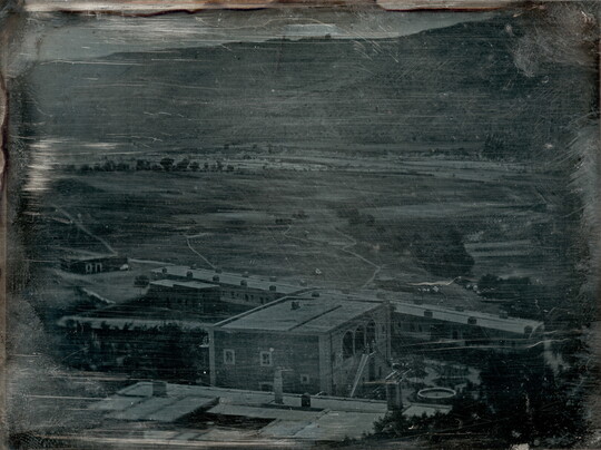 A black-and-white photograph, slightly obscured by surface marks, of a factory complex in front of a hillside.