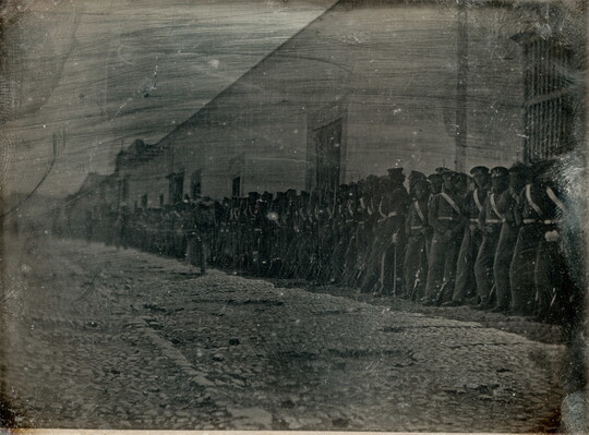 A dark, black-and-white photograph of a group of figures in military uniforms lined up against a building on a cobblestone street.
