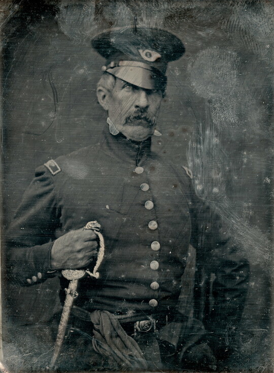 A black-and-white portrait of a White, male soldier in a dress uniform holding the hilt of a sword.