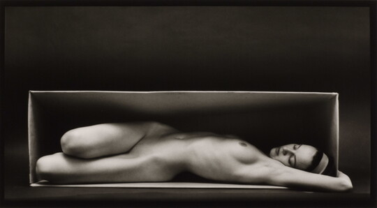 A black-and-white photograph of a nude, fair-skinned woman lying on her back, twisting toward the viewer, in a box.