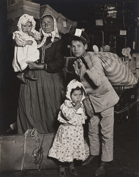 A black-and-white photograph of a light-skinned woman with three children of different ages, standing with suitcases and bags.