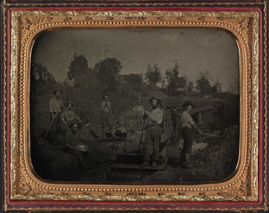 A black-and-white photograph in a gold frame of men posed with shovels and picks along a creek bed.