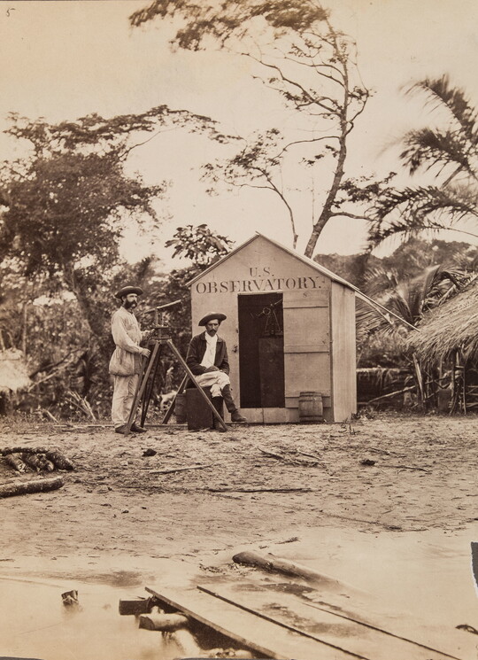 A black-and-white photograph of two men with a telescope standing in front of a small shack with "U.S. Observatory" above the doorway.
