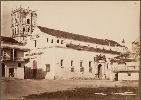 A sepia-toned photograph of dirt forecourt leading to a light-colored two-story structure and an ornate square tower in the background.