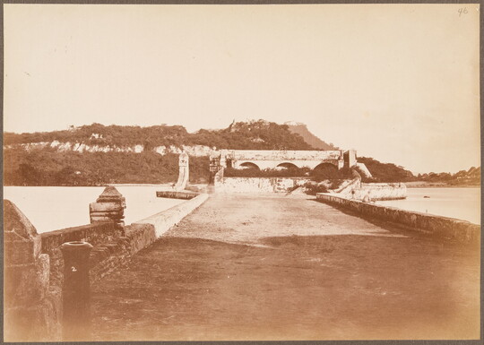 A sepia-toned photograph of a wide stone bridge leading across a river to arched structures on a rocky hillside.