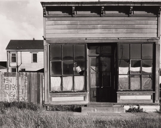 A black-and-white photograph of a wood storefront with cloth covering the windows and doors on the inside; overgrown grass lines the front walk.