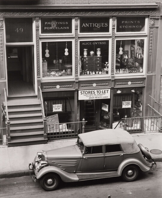 A black-and-white photograph of a vintage car parked on the street outside of a storefront advertising antiques.